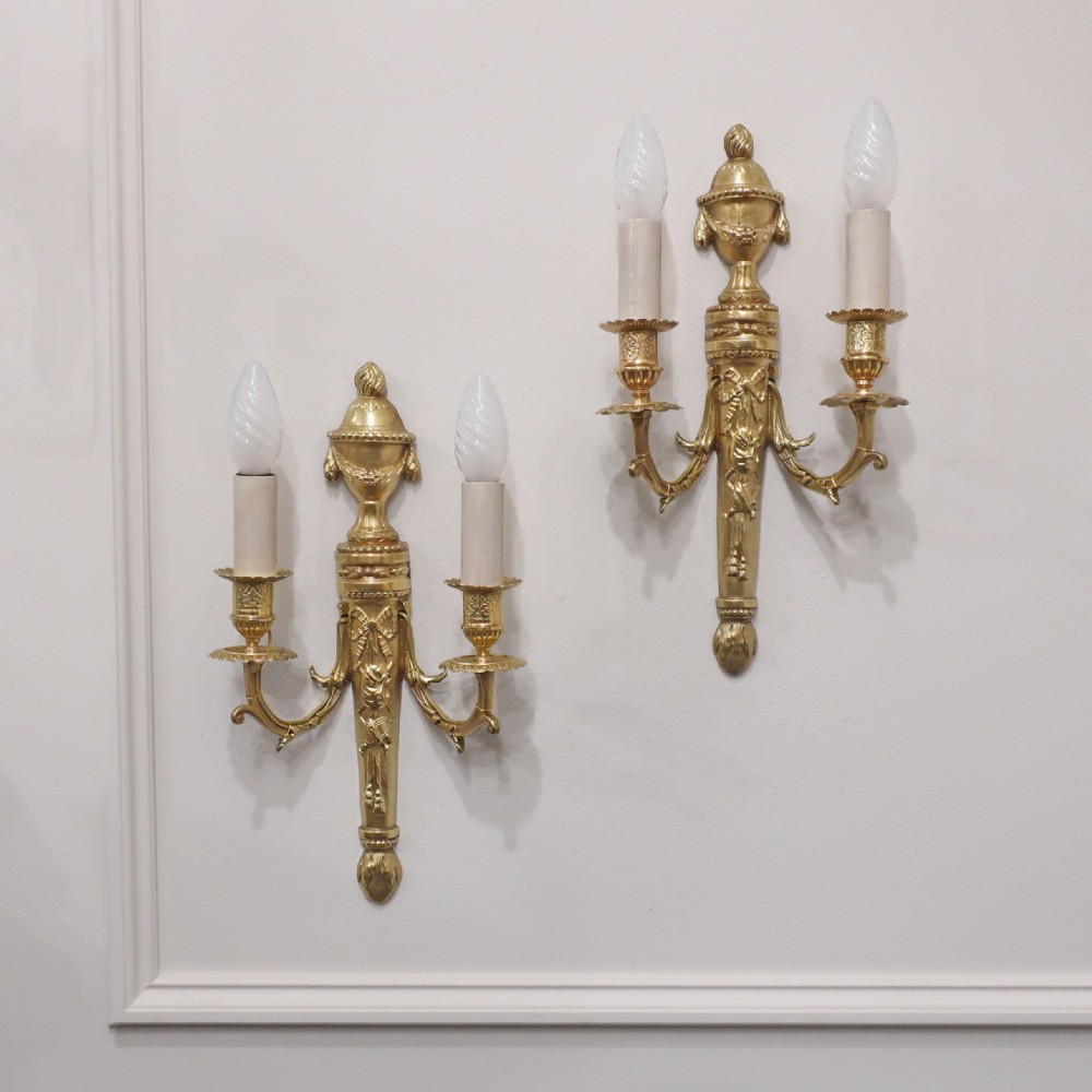 A pair of French Louis XVI style gilt bronze twin arm wall lights featuring classical urn finials and ribbon relief decorations.