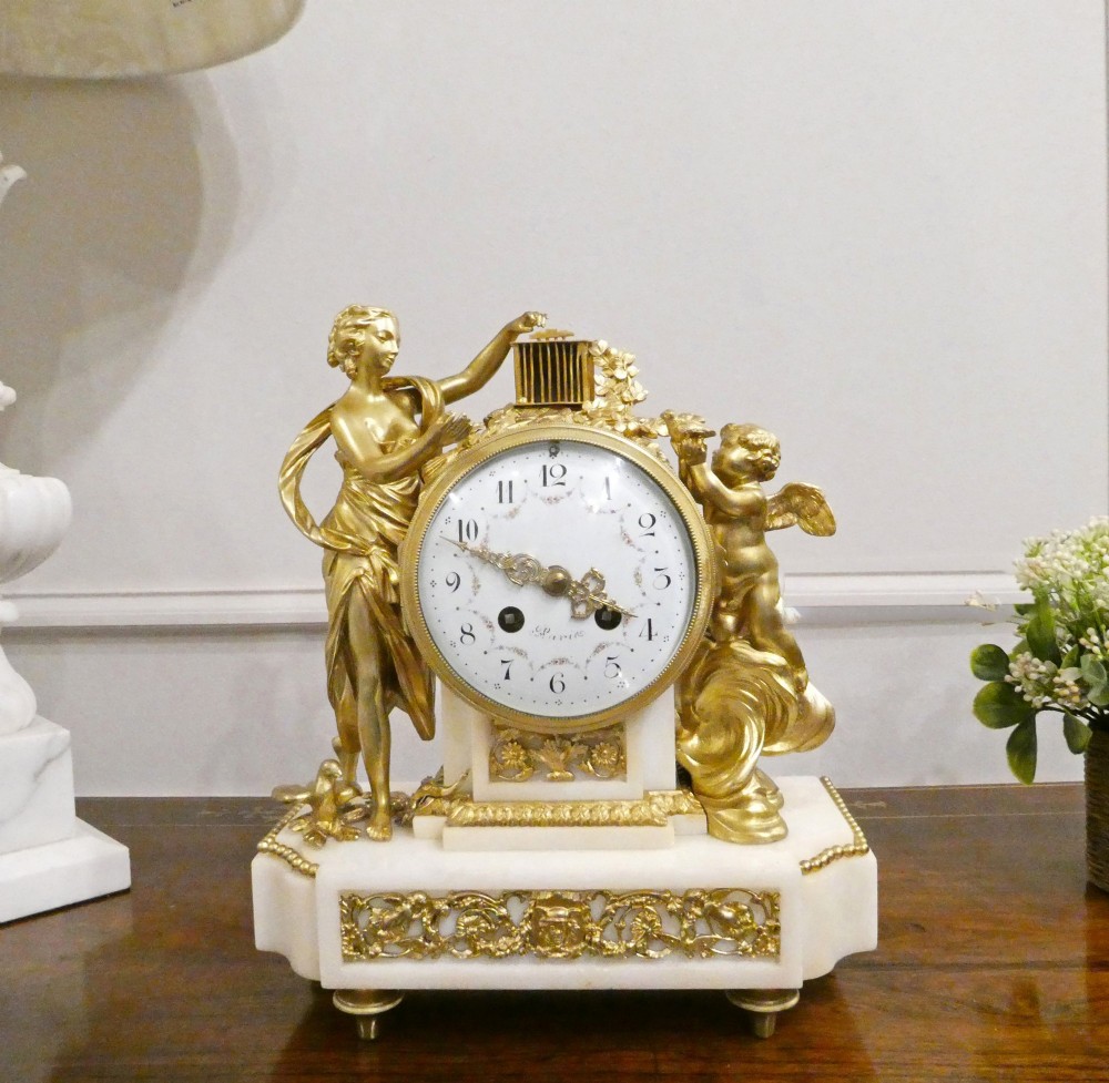 A French Louis XVI style gilt bronze and white marble mantel clock featuring classical female and winged cherub figures surrounding a white enamel dial.