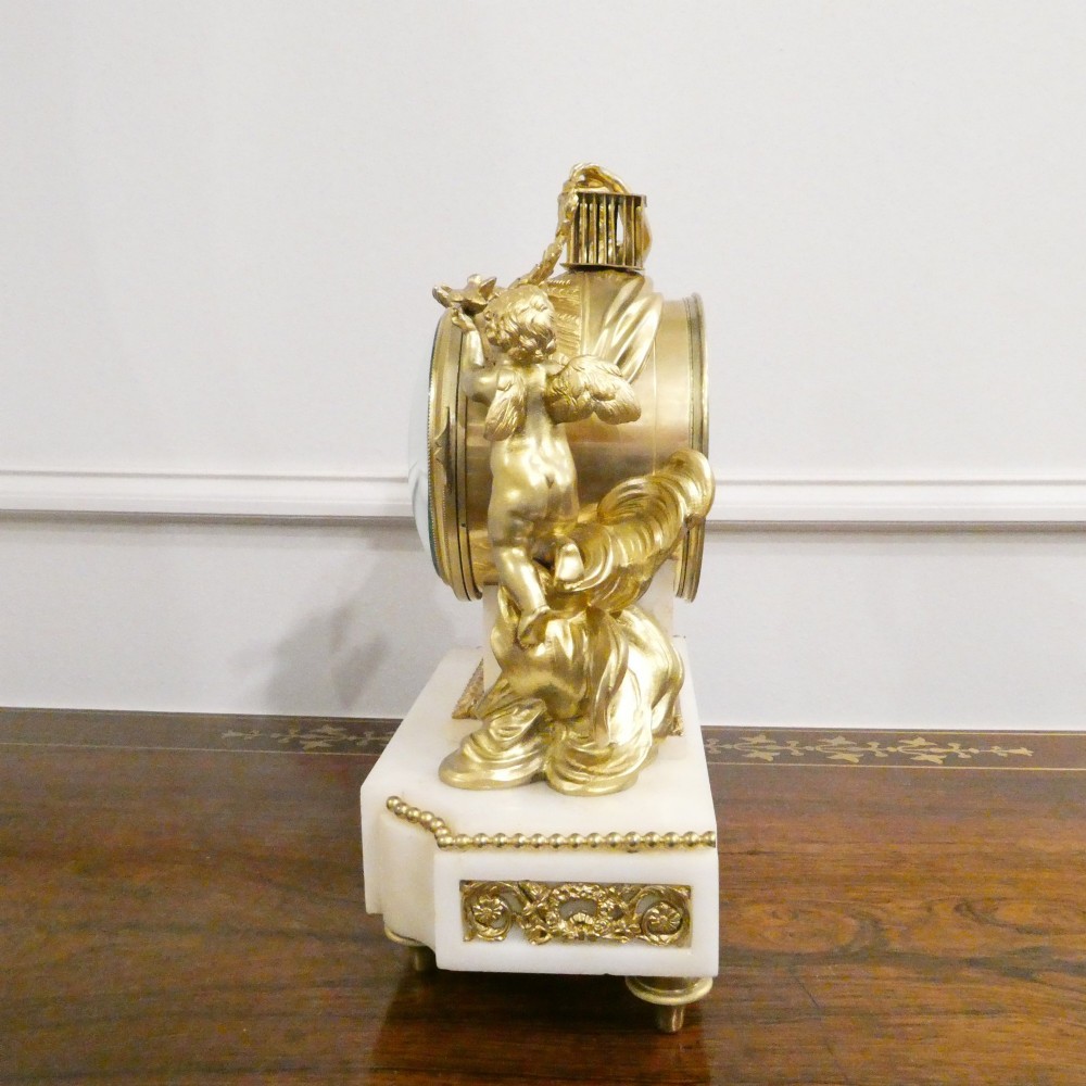A French Louis XVI style gilt bronze and white marble mantel clock featuring classical female and winged cherub figures surrounding a white enamel dial. View XII