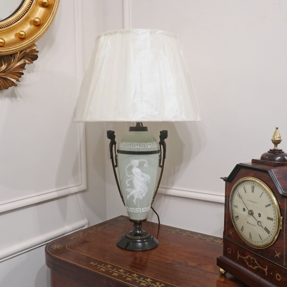 A French Neoclassical style table lamp featuring a pale green pate-sur-pate porcelain urn base, dark bronze side handles, and a pleated cream shade.