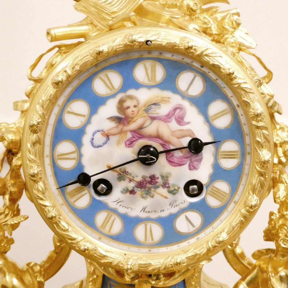 An ornate antique French gilt bronze mantel clock featuring decorative cherub figures, a recumbent goat sculpture, and blue porcelain panels painted with cherubs and floral motifs. View II