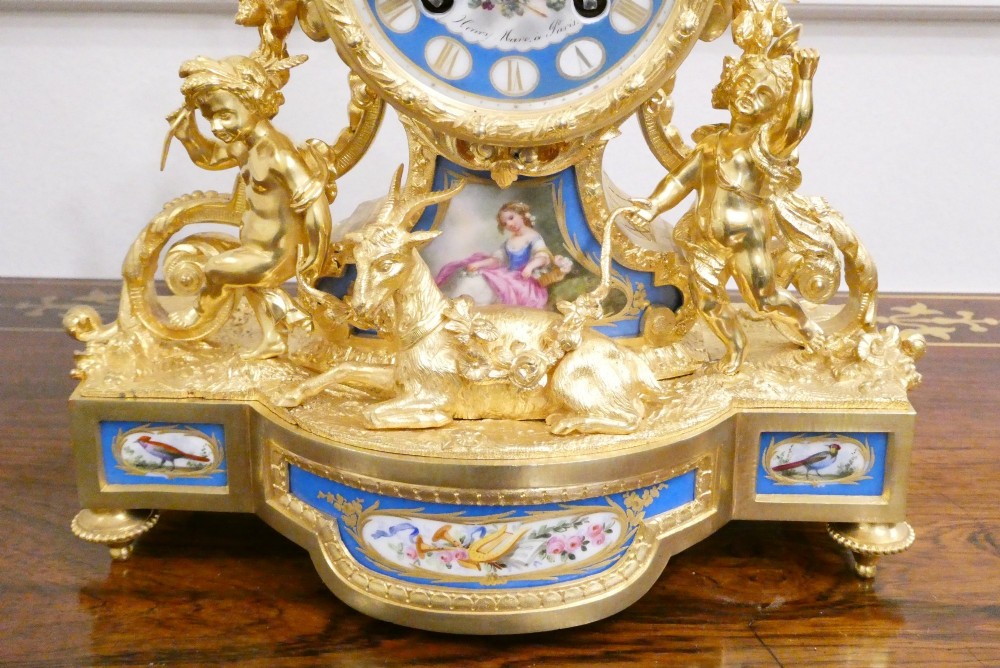 An ornate antique French gilt bronze mantel clock featuring decorative cherub figures, a recumbent goat sculpture, and blue porcelain panels painted with cherubs and floral motifs. View VI