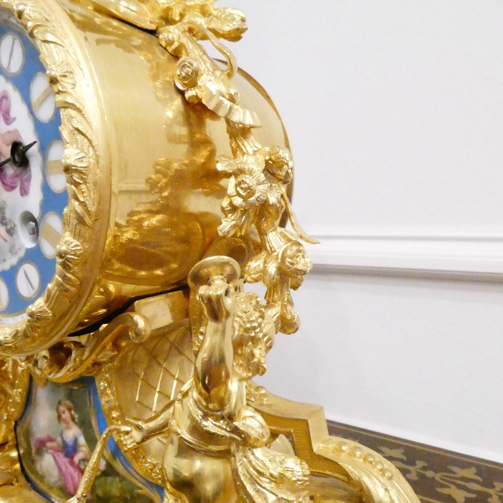 An ornate antique French gilt bronze mantel clock featuring decorative cherub figures, a recumbent goat sculpture, and blue porcelain panels painted with cherubs and floral motifs. View IX