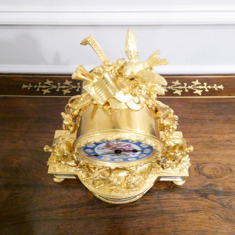An ornate antique French gilt bronze mantel clock featuring decorative cherub figures, a recumbent goat sculpture, and blue porcelain panels painted with cherubs and floral motifs. View X
