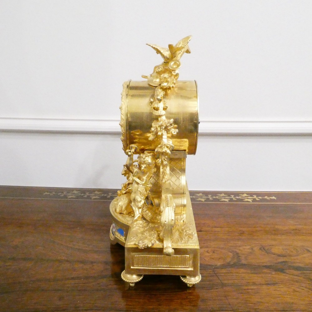 An ornate antique French gilt bronze mantel clock featuring decorative cherub figures, a recumbent goat sculpture, and blue porcelain panels painted with cherubs and floral motifs. View XI