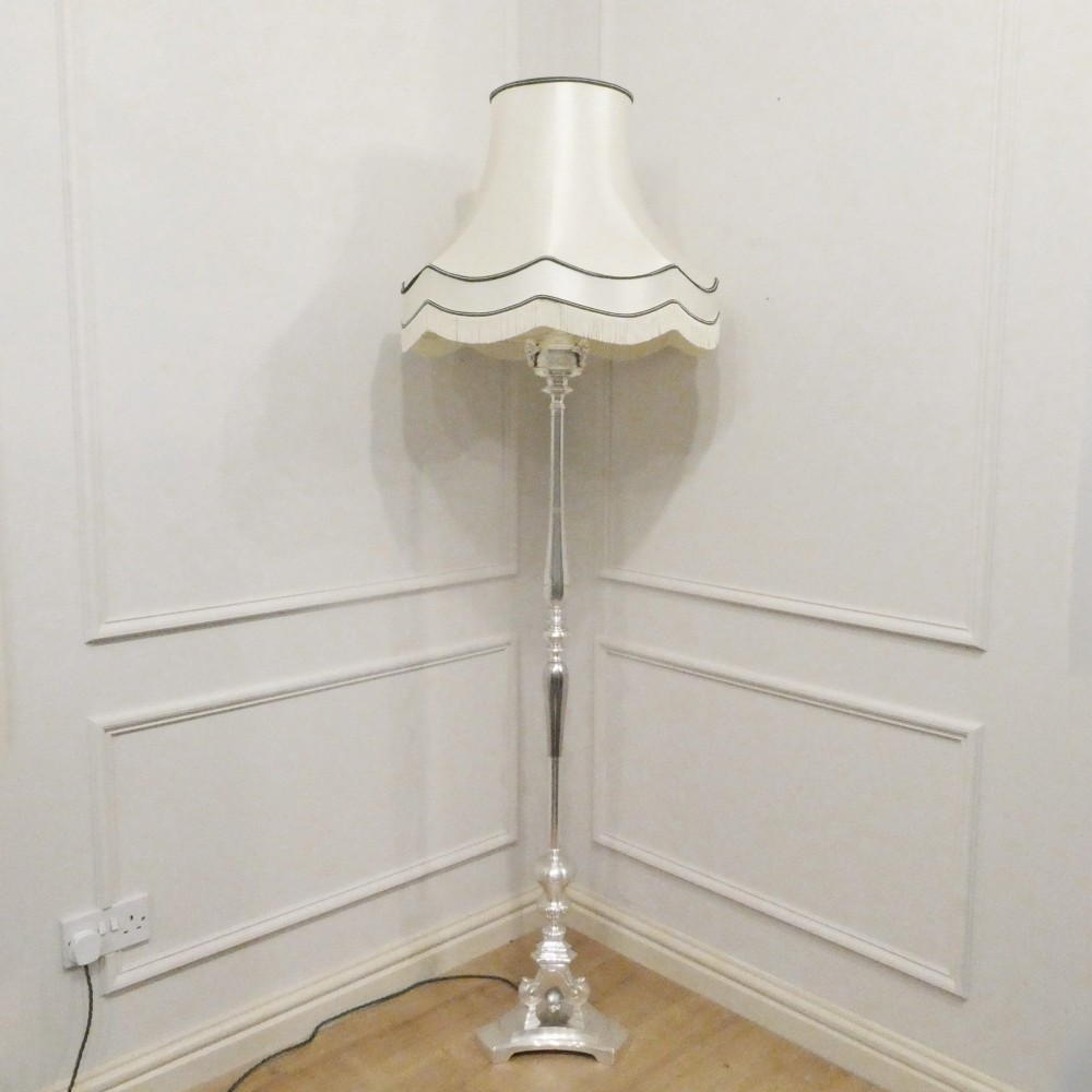 An early 20th-century silver-plated standard lamp featuring a turned column, cast base, and a cream scalloped lampshade with fringe detailing.