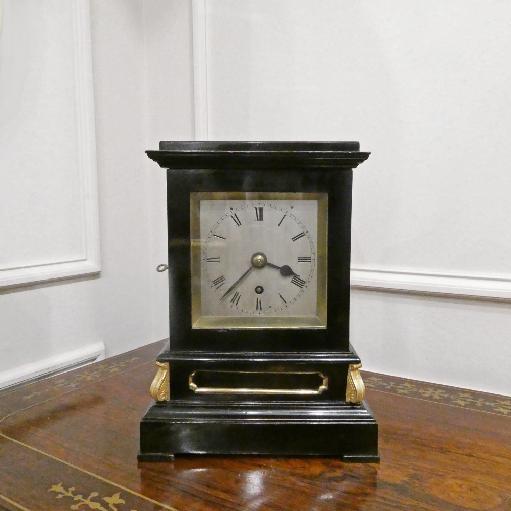 An early Victorian ebonised wooden library clock featuring a square dial with Roman numerals, side viewing apertures, and brass accents. View II