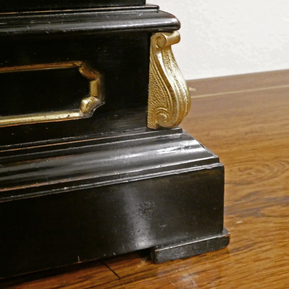 An early Victorian ebonised wooden library clock featuring a square dial with Roman numerals, side viewing apertures, and brass accents. View V