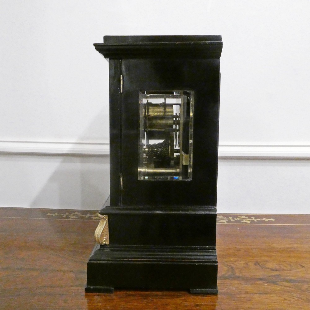 An early Victorian ebonised wooden library clock featuring a square dial with Roman numerals, side viewing apertures, and brass accents. View X