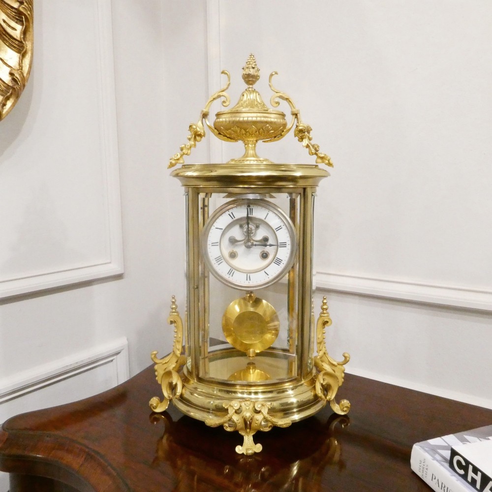 A French gilt bronze and brass four-glass mantel clock featuring an urn finial, visible pendulum mechanism, and white enamel dial with Roman numerals.