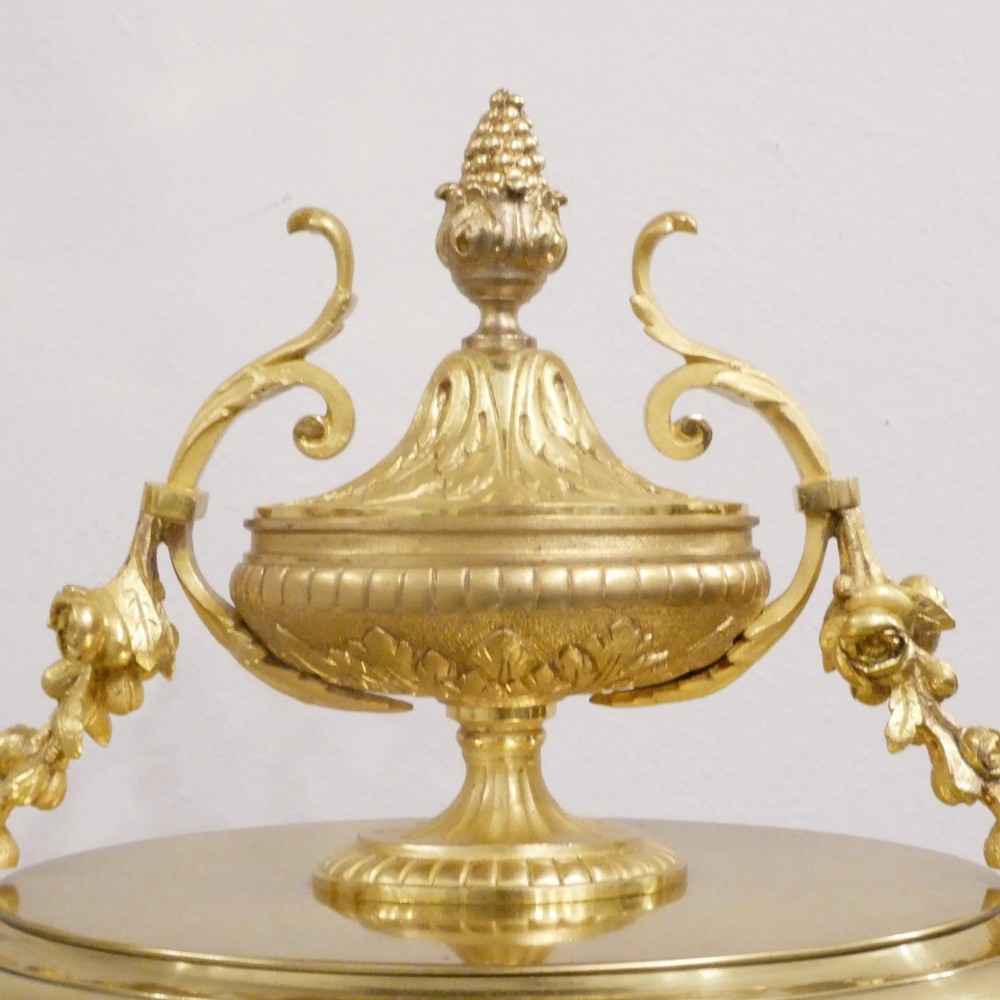 A French gilt bronze and brass four-glass mantel clock featuring an urn finial, visible pendulum mechanism, and white enamel dial with Roman numerals. View IX