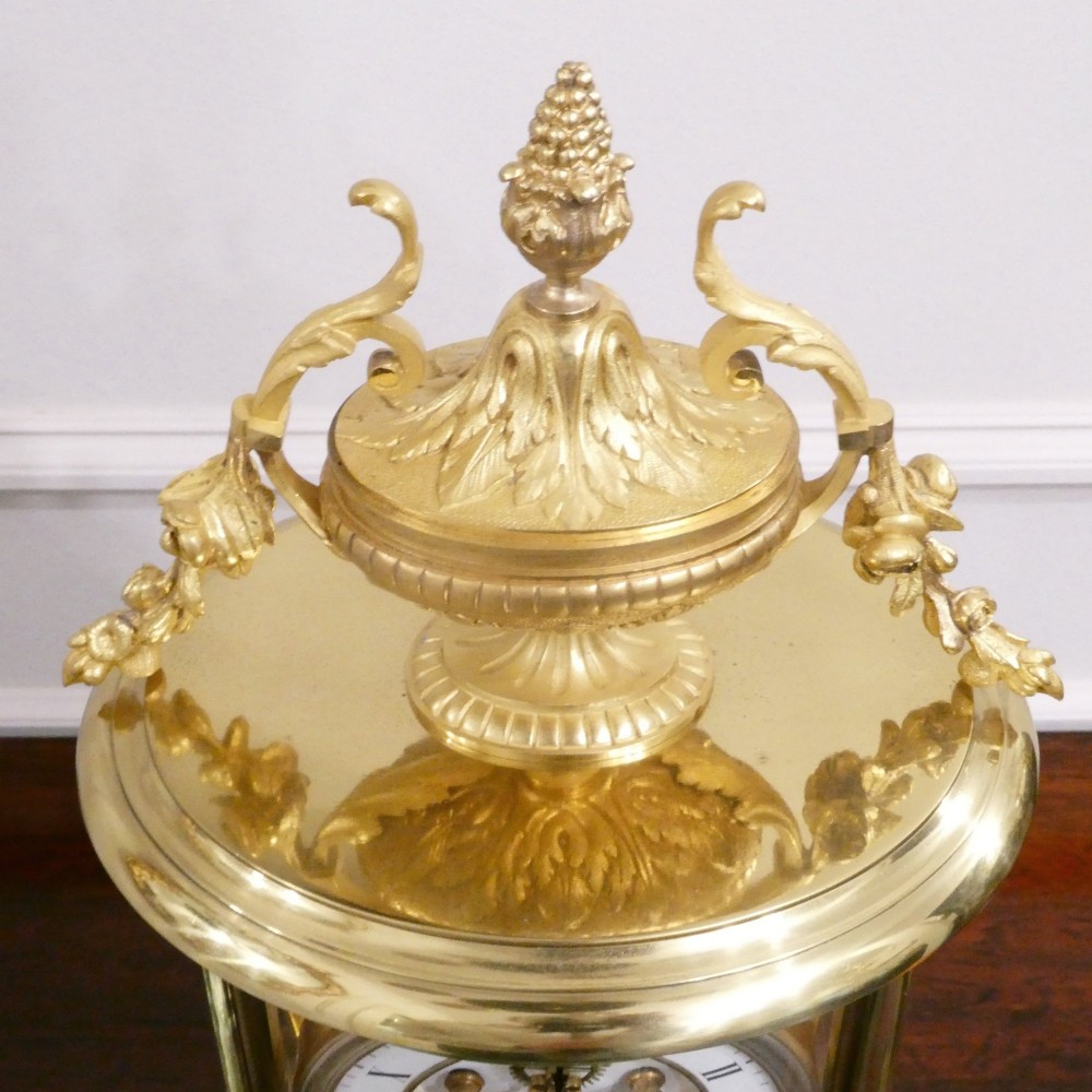 A French gilt bronze and brass four-glass mantel clock featuring an urn finial, visible pendulum mechanism, and white enamel dial with Roman numerals. View XI