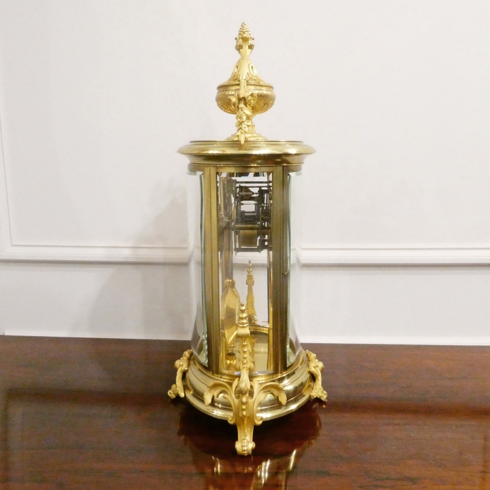 A French gilt bronze and brass four-glass mantel clock featuring an urn finial, visible pendulum mechanism, and white enamel dial with Roman numerals. View XII
