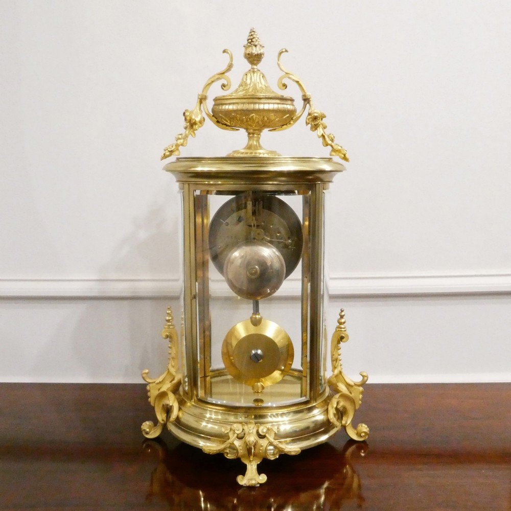 A French gilt bronze and brass four-glass mantel clock featuring an urn finial, visible pendulum mechanism, and white enamel dial with Roman numerals. View XIV