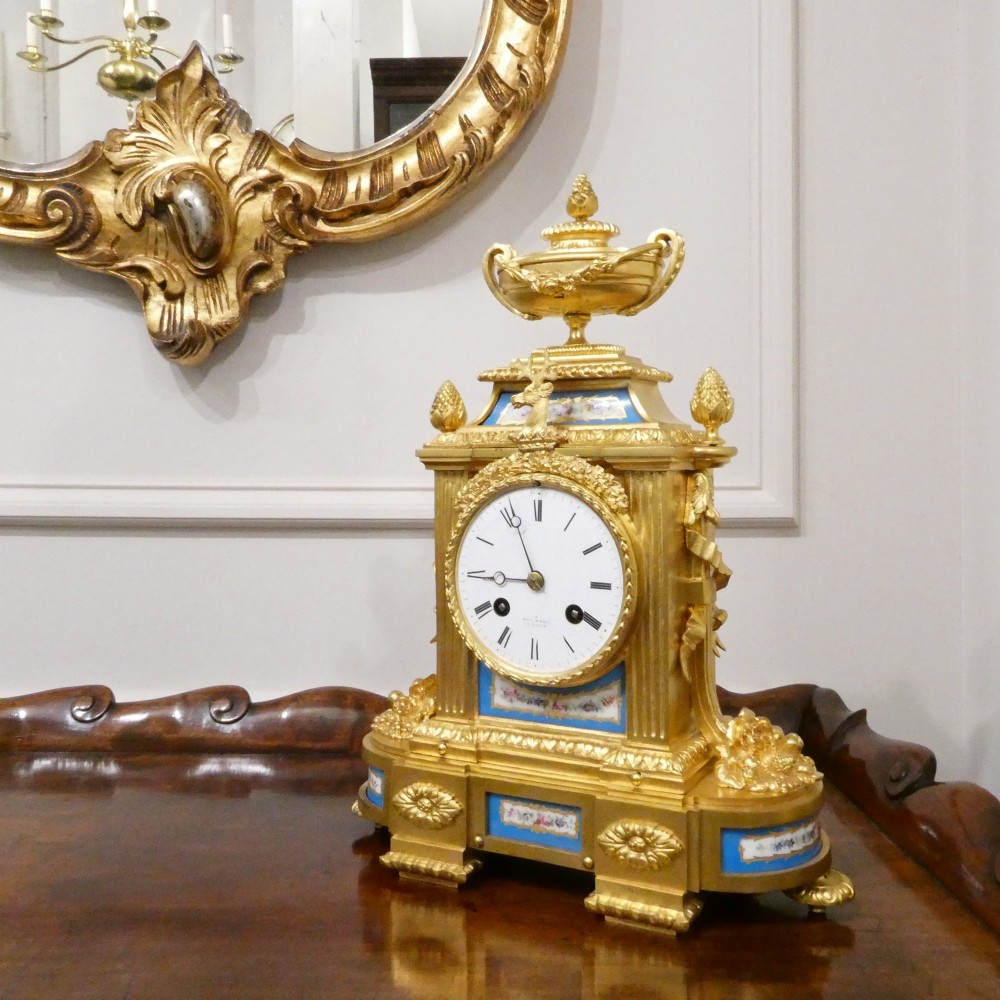 A French Louis XVI style gilt bronze mantel clock featuring a top urn finial, fluted side columns, and hand-painted blue porcelain panels around a white enamel dial. View II