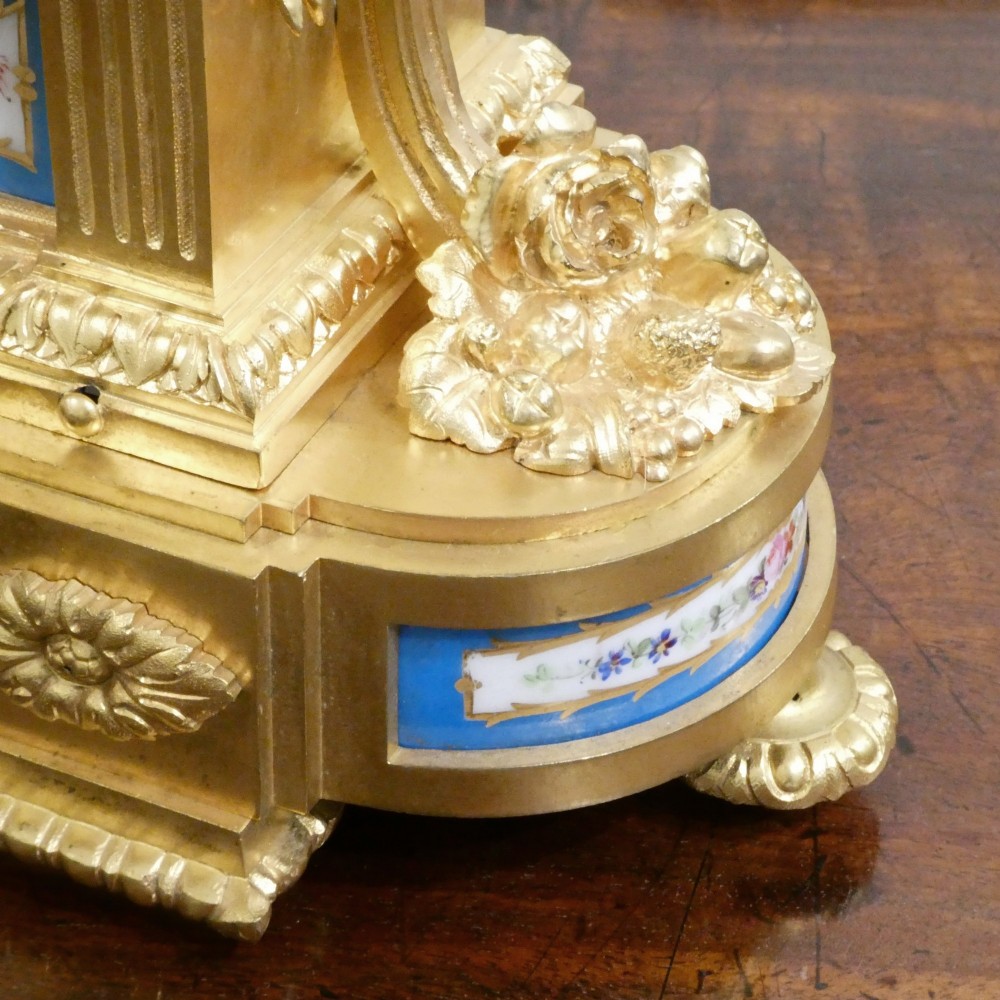 A French Louis XVI style gilt bronze mantel clock featuring a top urn finial, fluted side columns, and hand-painted blue porcelain panels around a white enamel dial. View VI