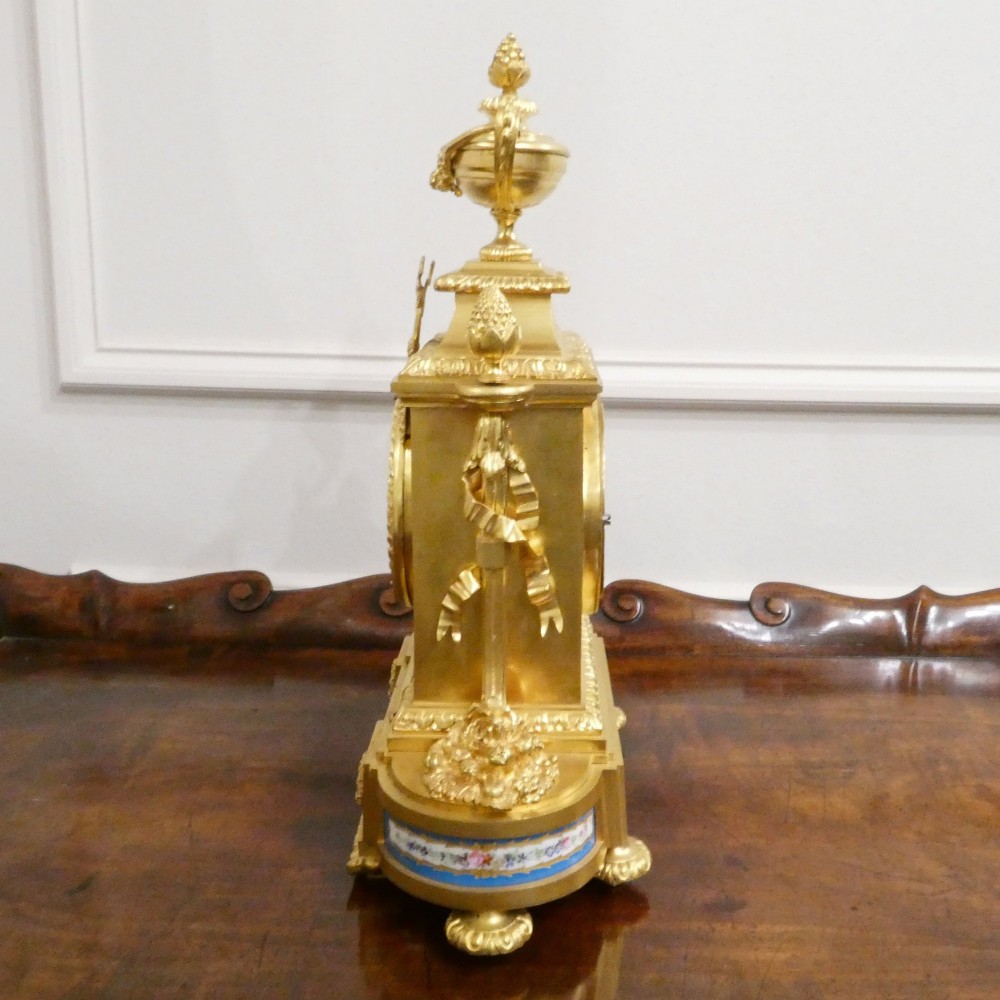 A French Louis XVI style gilt bronze mantel clock featuring a top urn finial, fluted side columns, and hand-painted blue porcelain panels around a white enamel dial. View XII
