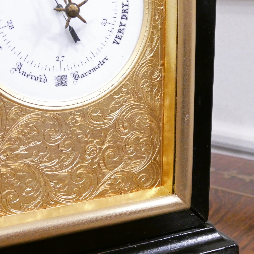 An Edwardian rectangular ebonised wooden mantel clock and barometer unit with twin white circular dials set into a scroll-engraved brass plate. View V