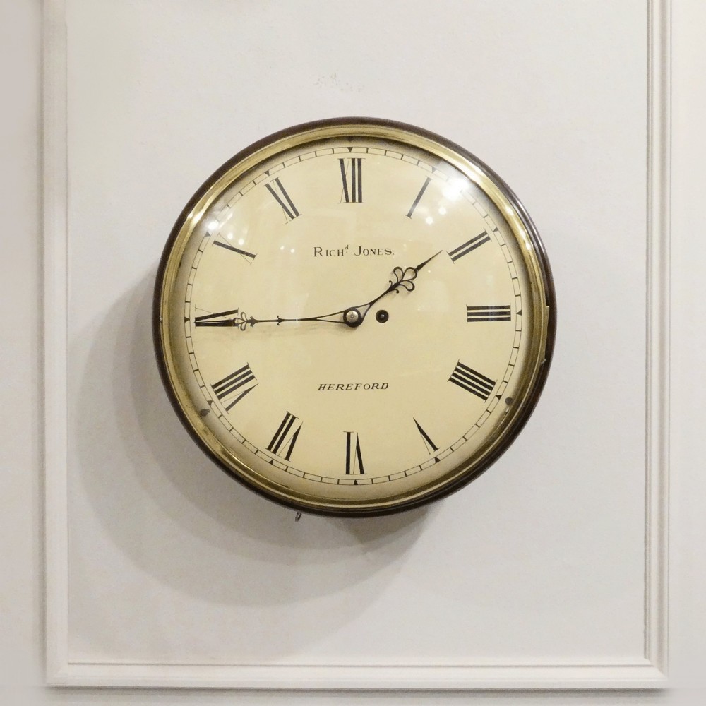A circular Georgian wall clock featuring a cream dial with black Roman numerals, blued steel hands, a polished brass bezel, and a dark mahogany wood surround.