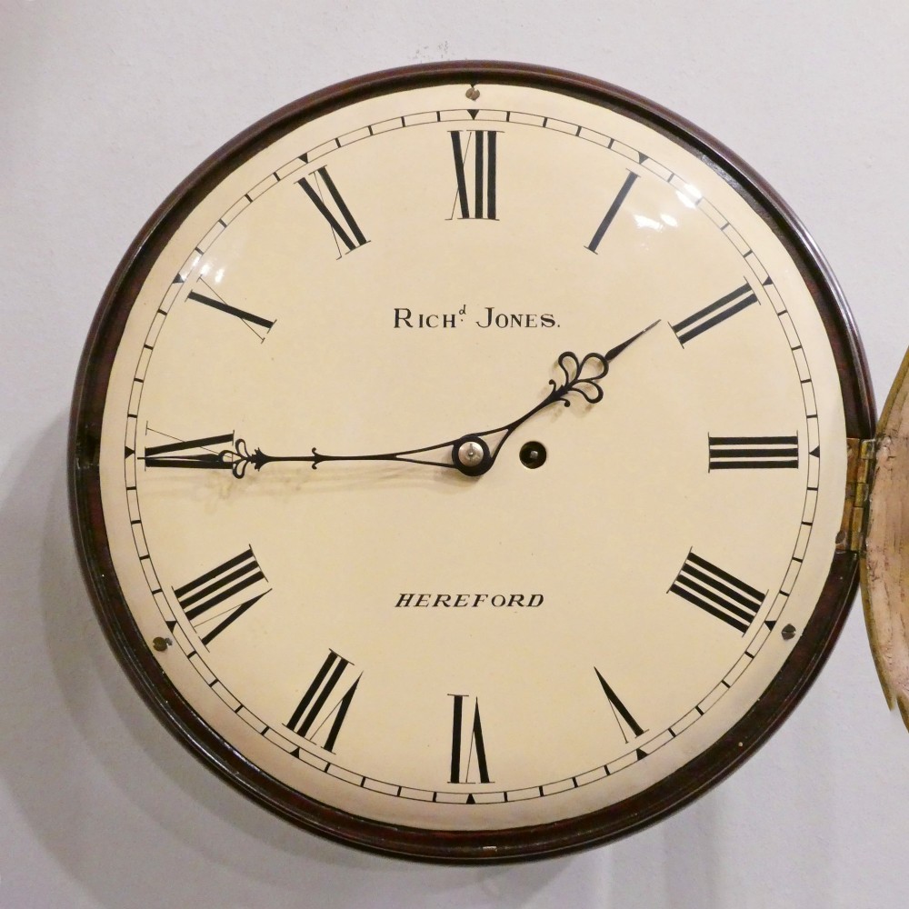 A circular Georgian wall clock featuring a cream dial with black Roman numerals, blued steel hands, a polished brass bezel, and a dark mahogany wood surround. View III