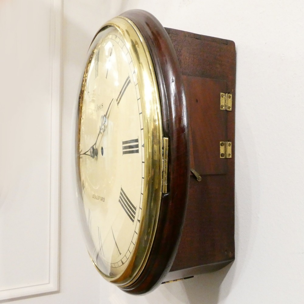 A circular Georgian wall clock featuring a cream dial with black Roman numerals, blued steel hands, a polished brass bezel, and a dark mahogany wood surround. View V