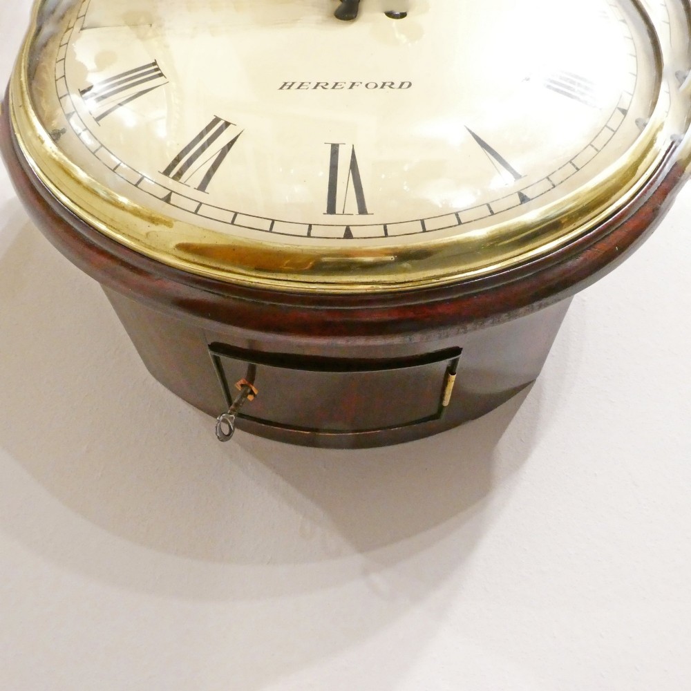A circular Georgian wall clock featuring a cream dial with black Roman numerals, blued steel hands, a polished brass bezel, and a dark mahogany wood surround. View VI