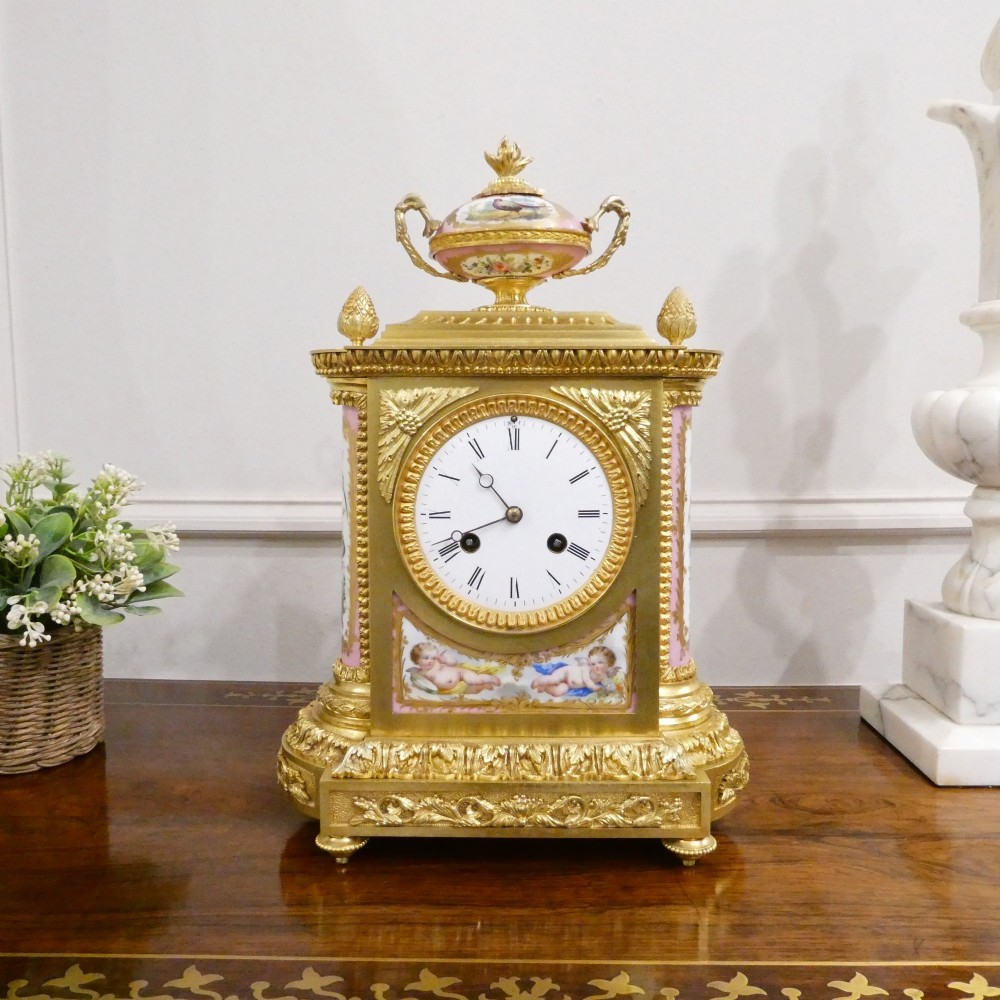 An antique French gilt bronze mantel clock featuring hand-painted pink porcelain panels, a porcelain urn finial, and an enamel dial with Roman numerals.