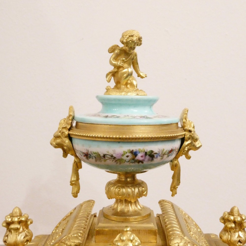 A French 19th-century gilt bronze mantel clock featuring light blue hand-painted porcelain panels, sculpted putti, an urn finial, and an enamel Roman numeral dial. View X