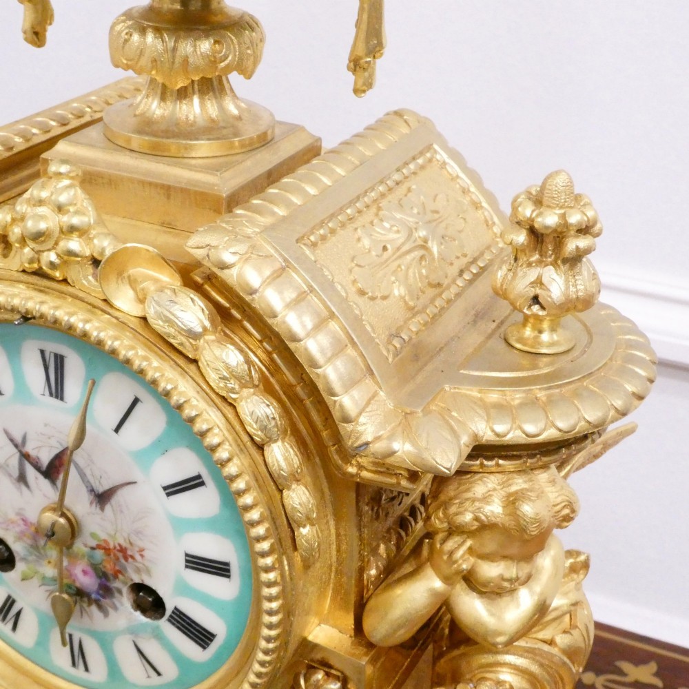 A French 19th-century gilt bronze mantel clock featuring light blue hand-painted porcelain panels, sculpted putti, an urn finial, and an enamel Roman numeral dial. View XII