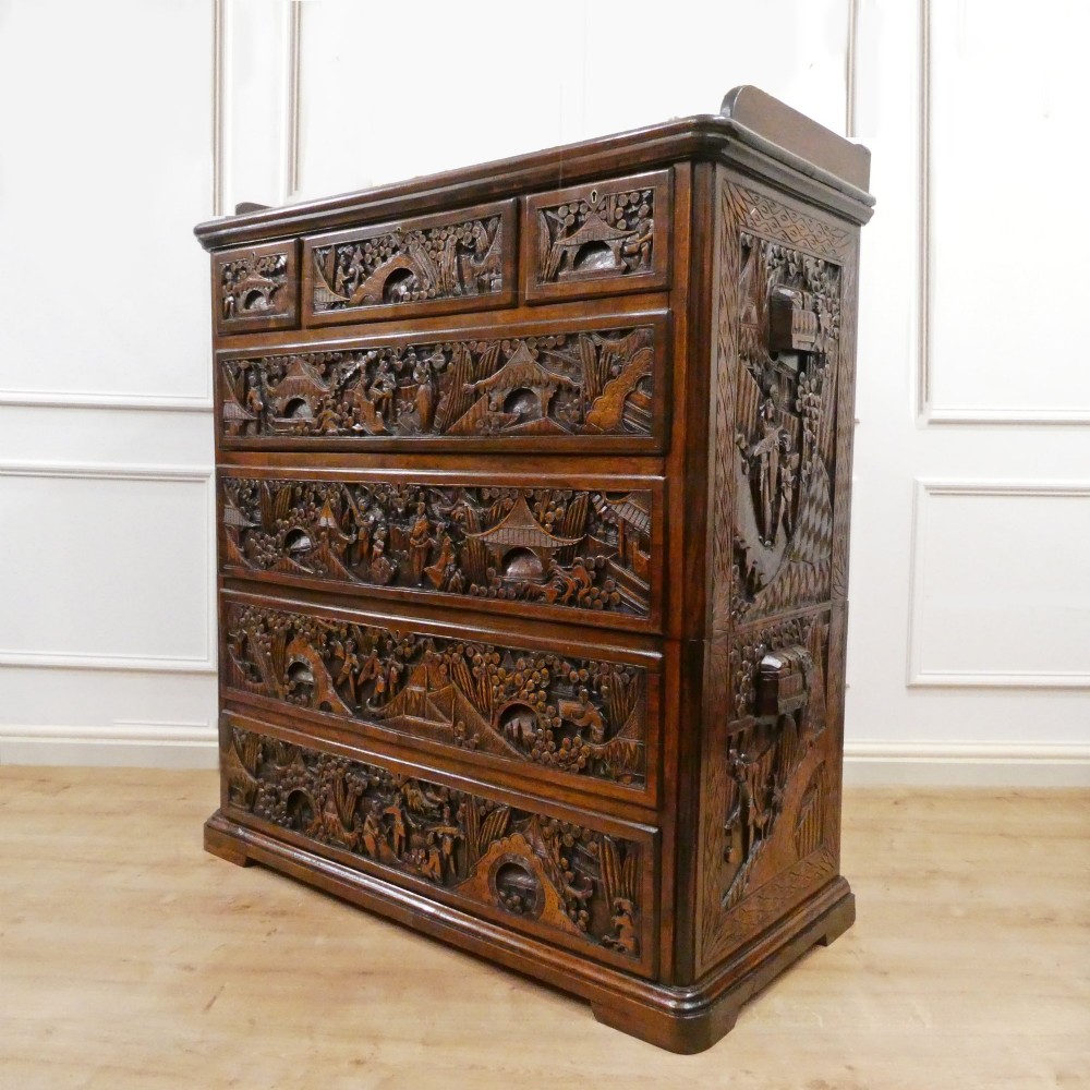 A tall early twentieth-century camphor wood chest of drawers featuring elaborate relief-carved oriental landscapes on the drawer fronts and side panels.