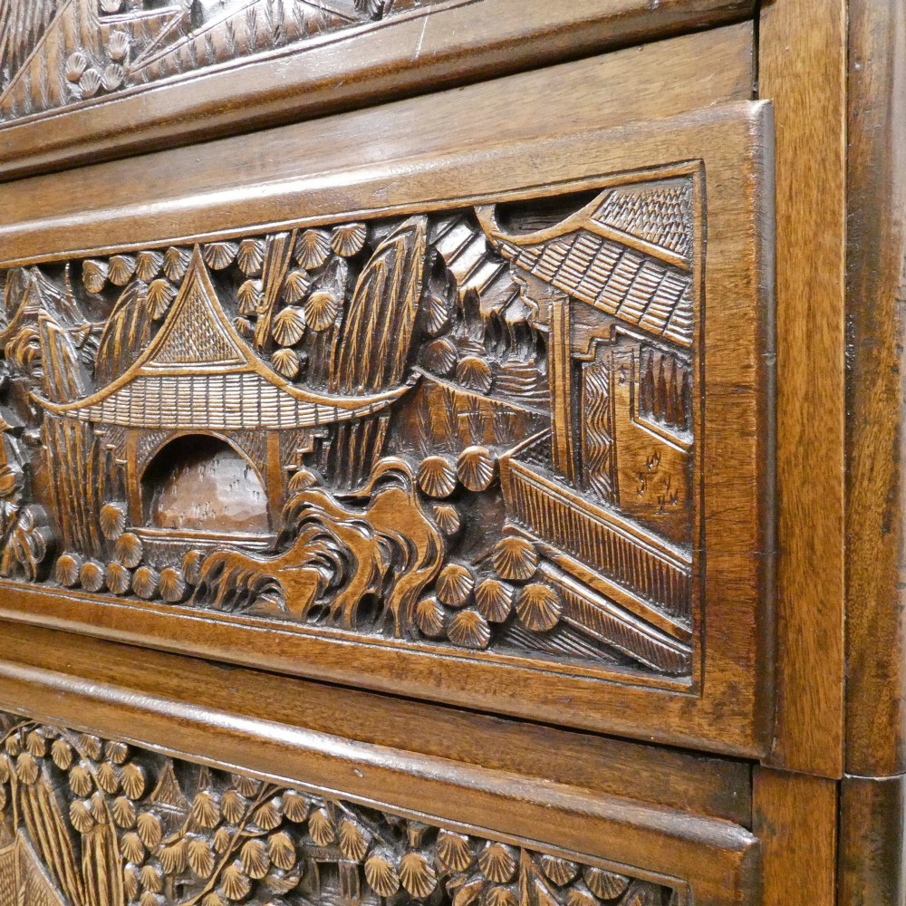 A tall early twentieth-century camphor wood chest of drawers featuring elaborate relief-carved oriental landscapes on the drawer fronts and side panels. View V