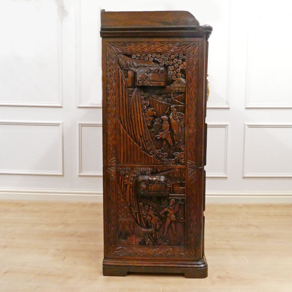 A tall early twentieth-century camphor wood chest of drawers featuring elaborate relief-carved oriental landscapes on the drawer fronts and side panels. View XII