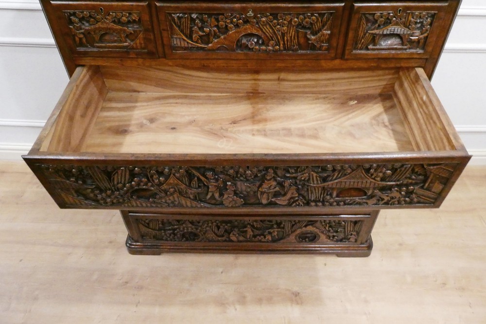 A tall early twentieth-century camphor wood chest of drawers featuring elaborate relief-carved oriental landscapes on the drawer fronts and side panels. View XV