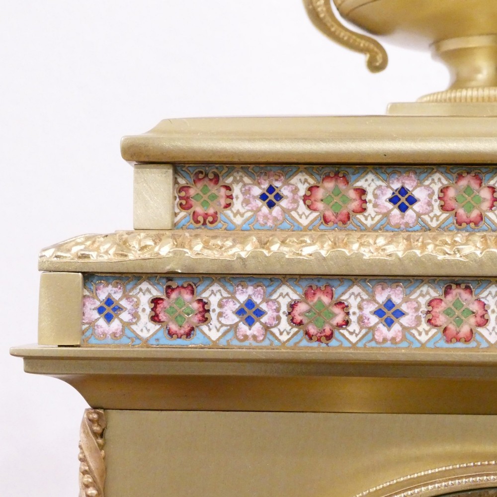 A nineteenth-century French gilt brass mantel clock featuring colourful champleve enamel geometric patterns, flanked by two matching decorative urns. View V