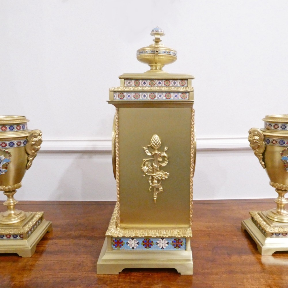 A nineteenth-century French gilt brass mantel clock featuring colourful champleve enamel geometric patterns, flanked by two matching decorative urns. View VIII