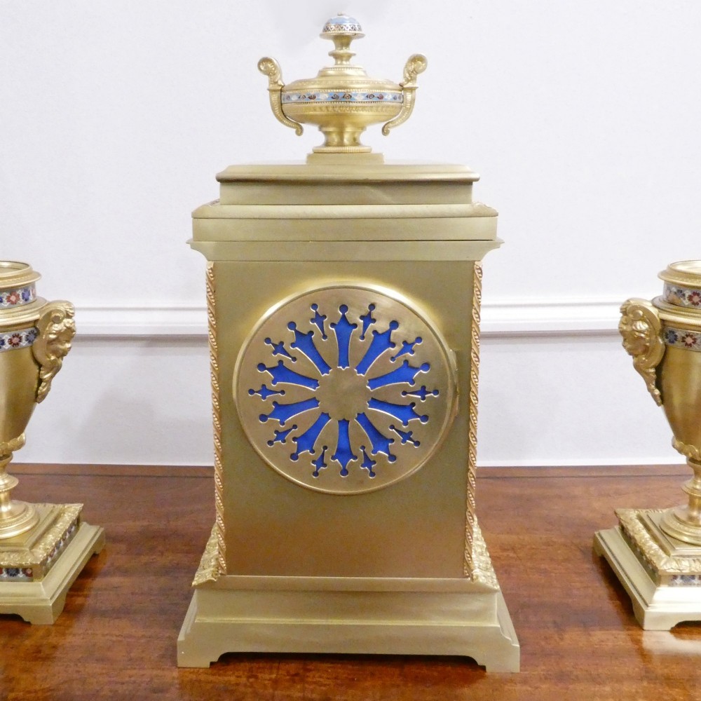 A nineteenth-century French gilt brass mantel clock featuring colourful champleve enamel geometric patterns, flanked by two matching decorative urns. View X
