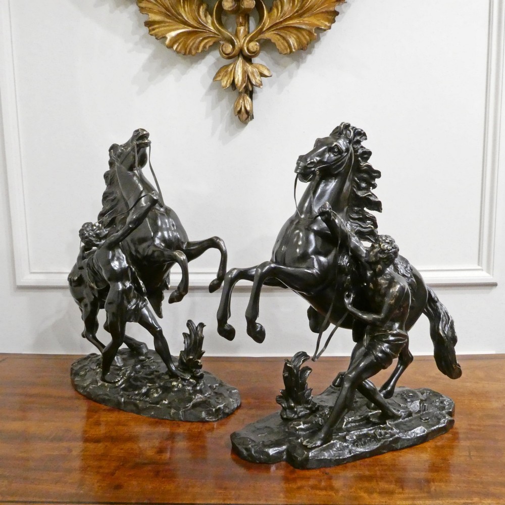A pair of patinated dark bronze sculptures depicting rearing horses held by grooms on naturalistic bases.