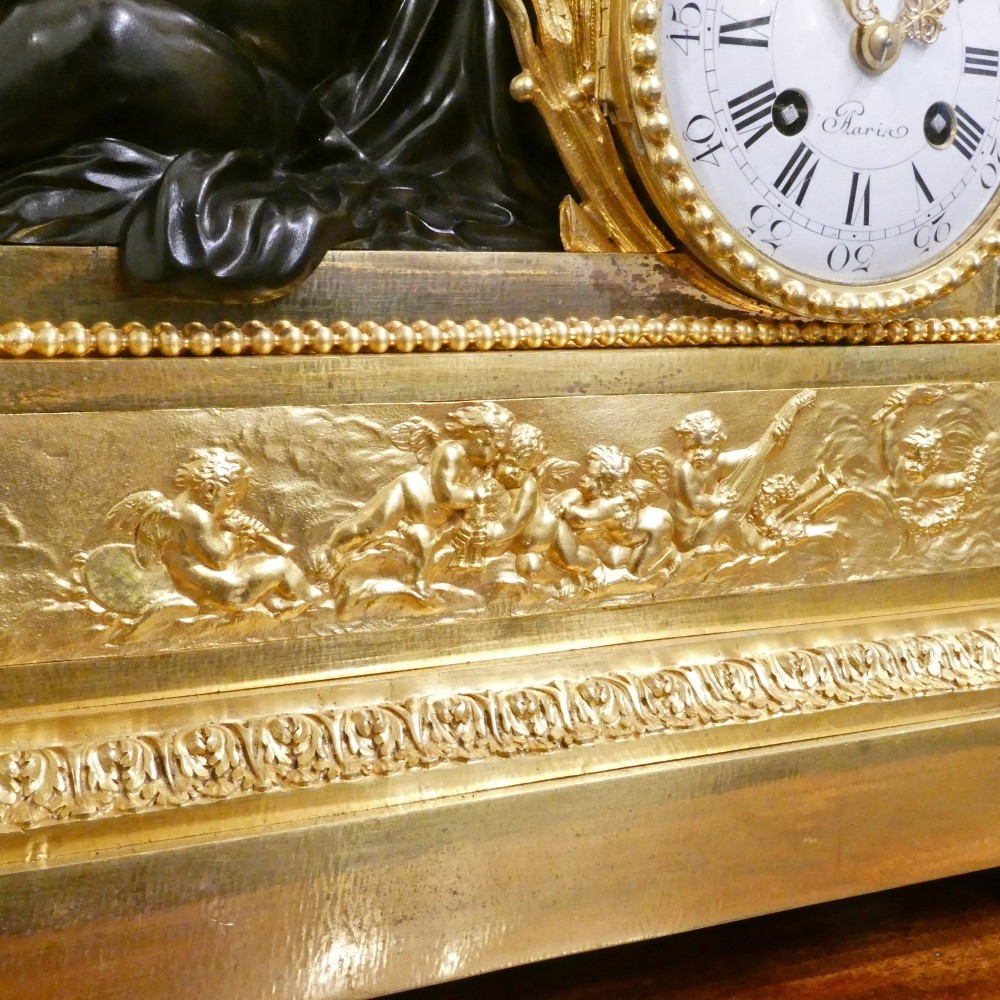 A 19th-century French gilt bronze mantel clock featuring a dark patinated bronze cherub seated beside a circular enamel dial atop a gilt relief base. View IX