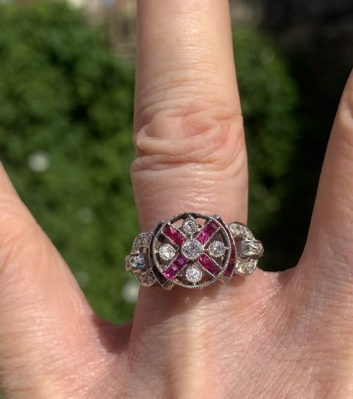 fig 1. An Art Deco ring featuring a central round diamond encircled by geometric cross-shaped calibre-cut rubies and additional diamond accents set in white metal.