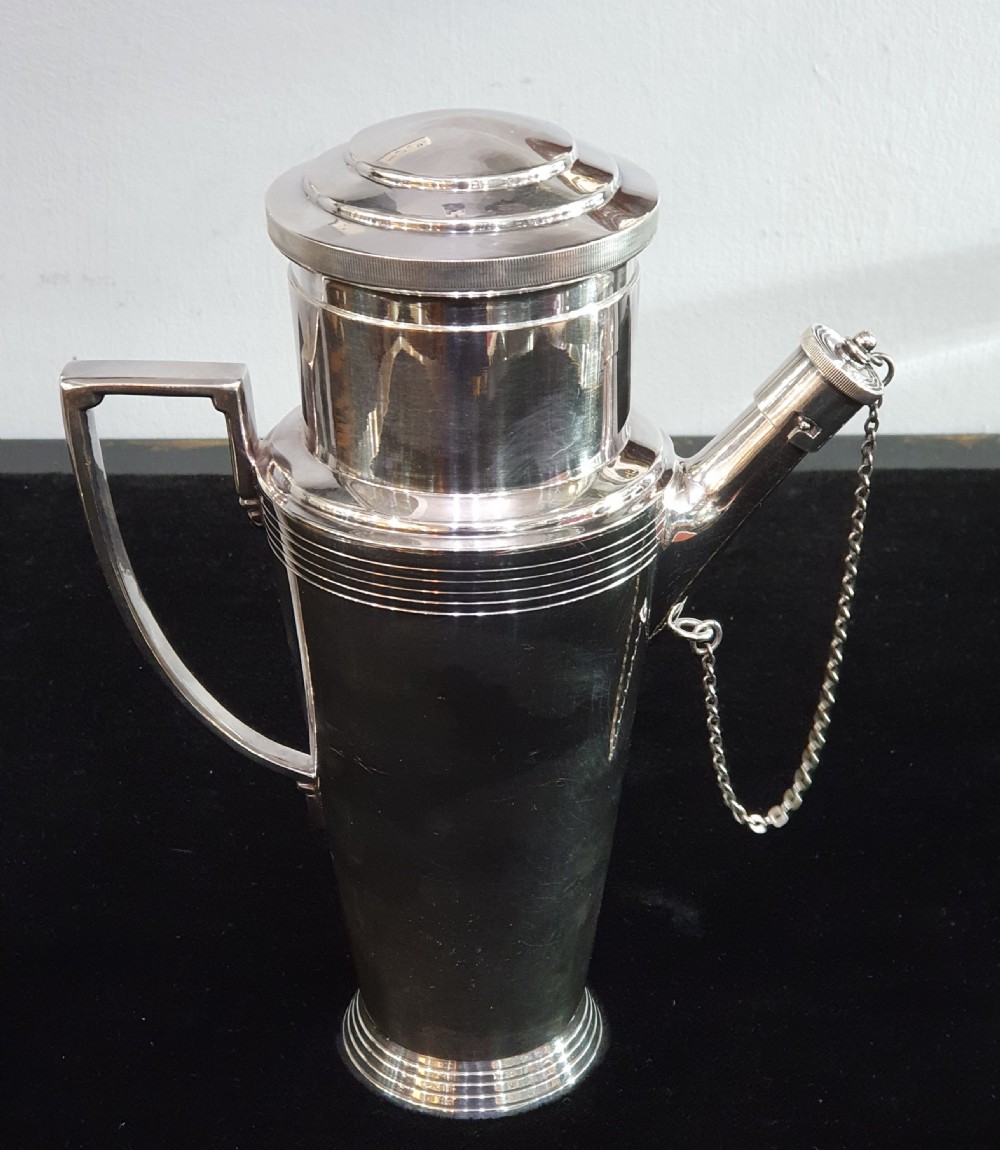 An Art Deco silver-plated cocktail shaker featuring a tapered conical body with turned concentric bands, an angular handle, and a spout with a chained cap stopper. View X