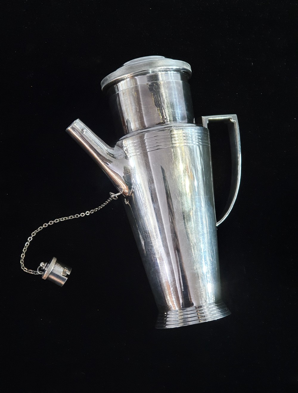 An Art Deco silver-plated cocktail shaker featuring a tapered conical body with turned concentric bands, an angular handle, and a spout with a chained cap stopper. View VI
