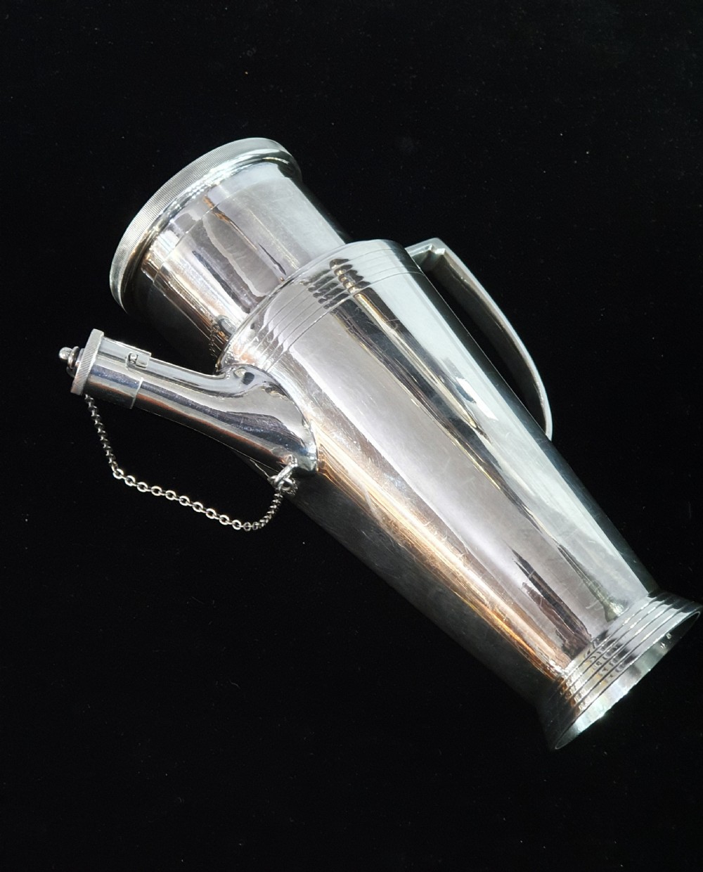 An Art Deco silver-plated cocktail shaker featuring a tapered conical body with turned concentric bands, an angular handle, and a spout with a chained cap stopper. View VII