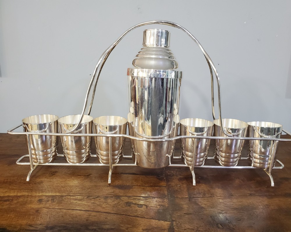 An Art Deco silver-plated cocktail set featuring a central shaker, six matching beakers, and a fitted metal carrying rack with a high arching handle.