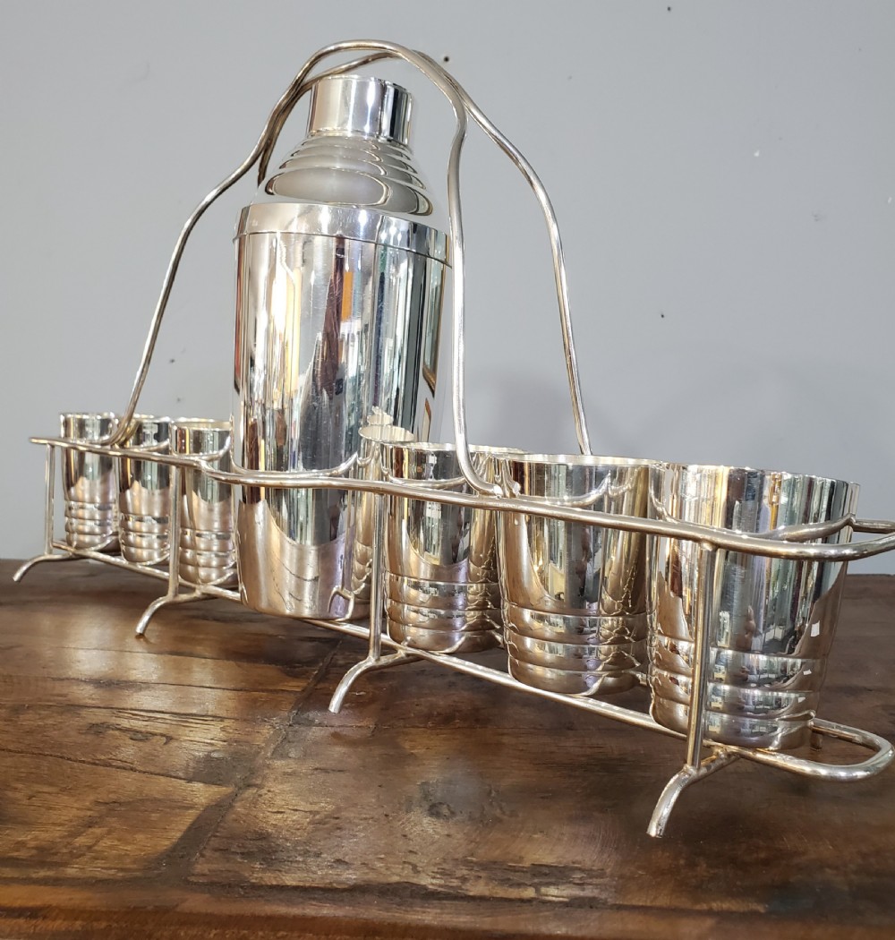 An Art Deco silver-plated cocktail set featuring a central shaker, six matching beakers, and a fitted metal carrying rack with a high arching handle. View II