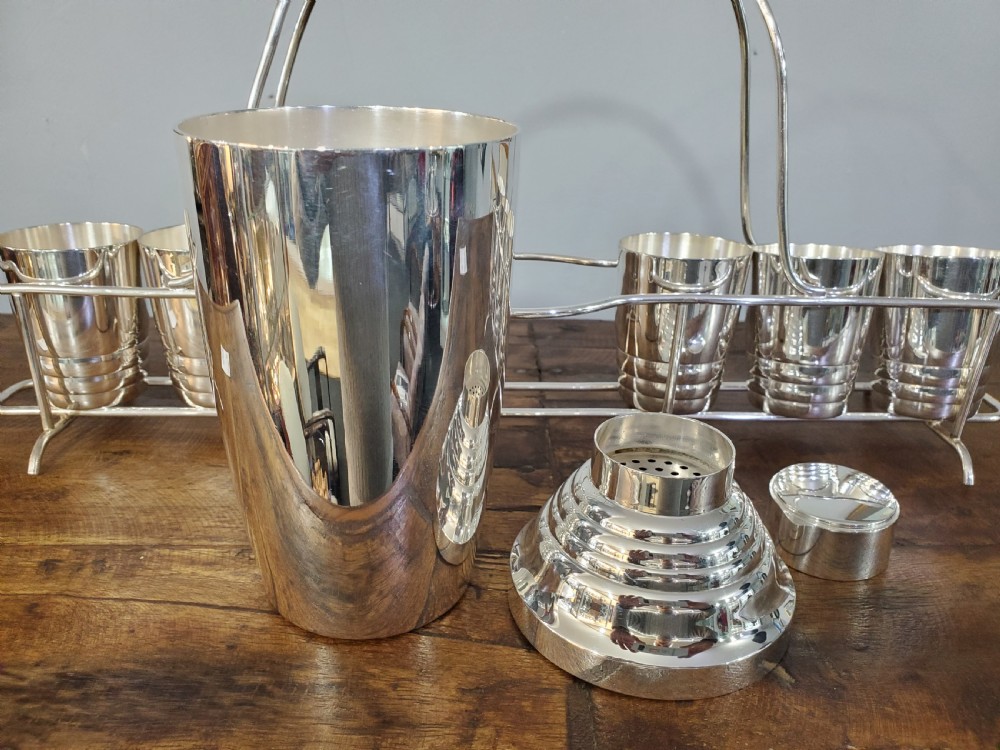 An Art Deco silver-plated cocktail set featuring a central shaker, six matching beakers, and a fitted metal carrying rack with a high arching handle. View IV