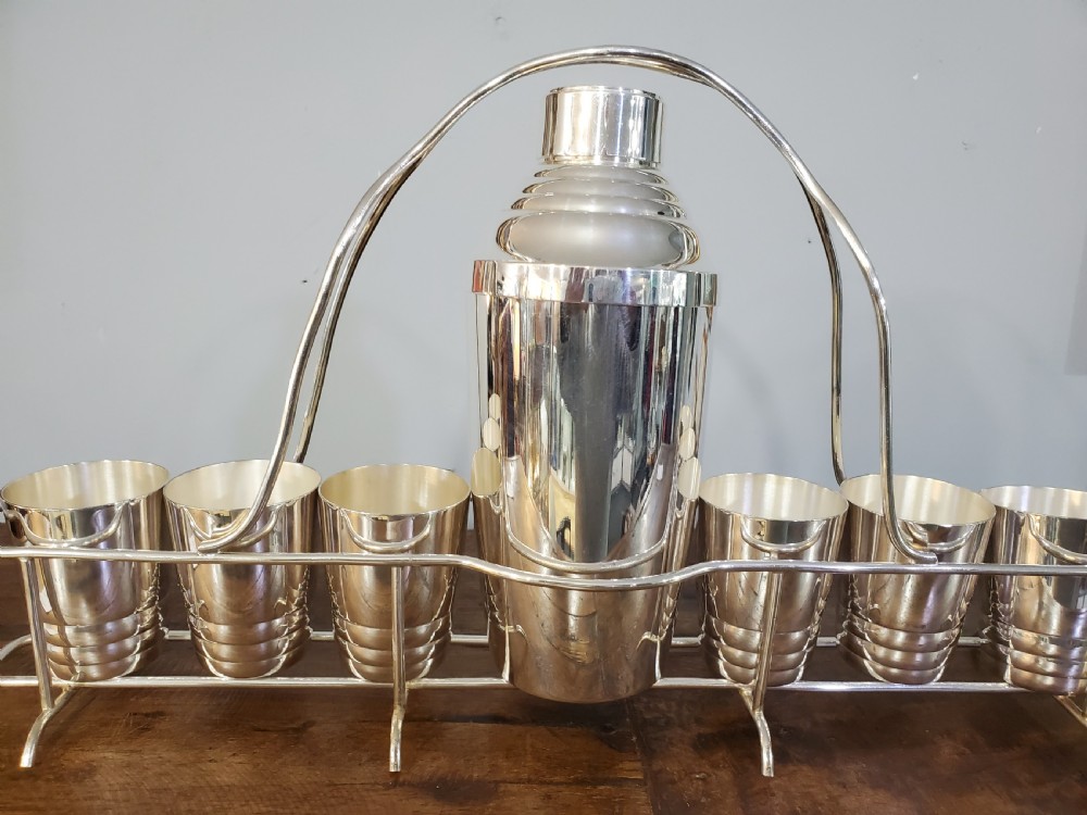 An Art Deco silver-plated cocktail set featuring a central shaker, six matching beakers, and a fitted metal carrying rack with a high arching handle. View VI