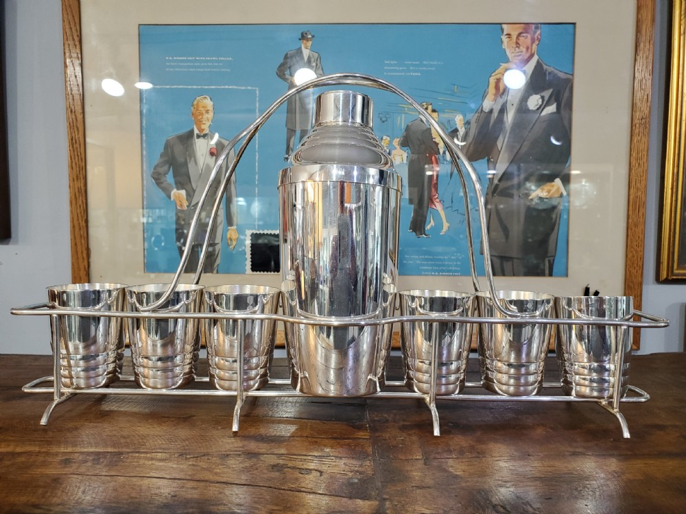 An Art Deco silver-plated cocktail set featuring a central shaker, six matching beakers, and a fitted metal carrying rack with a high arching handle. View VII