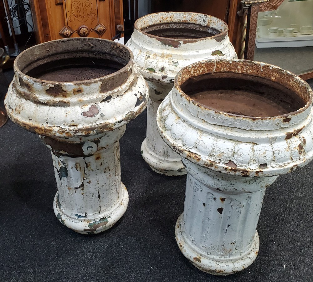 A trio of Victorian cast iron garden urns with fluted pedestals and flanged rims displaying chipped white paint and rust oxidisation.
