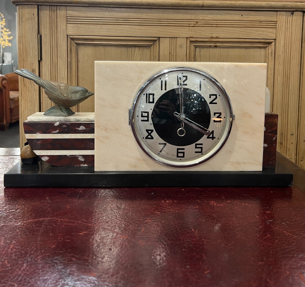 An Art Deco mantel clock featuring a cream marble face slab, contrasting dark striped marble accents, a metal bird statuette, and a round dial with bold Arabic numerals.