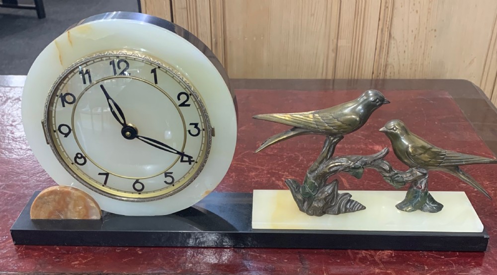 A French Art Deco mantel clock with a circular cream onyx timepiece mounted on a black marble base beside a metal bird sculpture.
